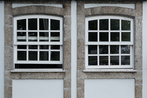 Two adjacent windows with white frames and multiple panes are set in a stone wall. Each window features a row of square panes at the top and rectangular panes below. The wall around the windows is constructed from textured gray stone with white borders. Different shades of green from trees are visible through the glass panes.