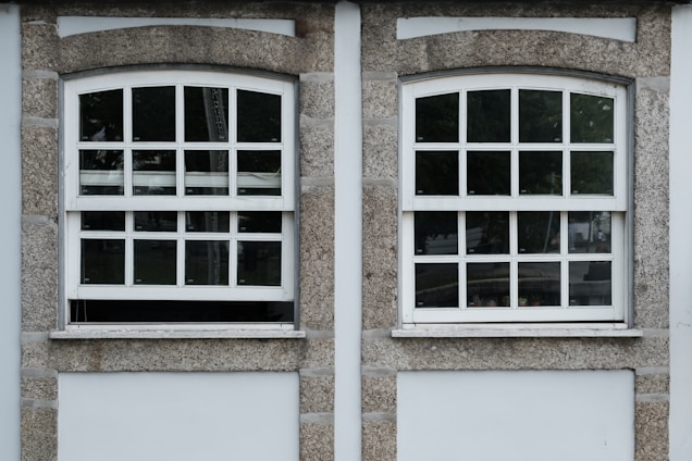 Two adjacent windows with white frames and multiple panes are set in a stone wall. Each window features a row of square panes at the top and rectangular panes below. The wall around the windows is constructed from textured gray stone with white borders. Different shades of green from trees are visible through the glass panes.