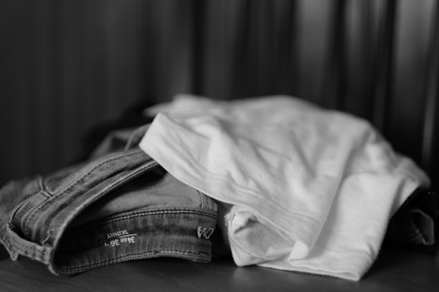 A neatly folded stack of clothing items including jeans and a white t-shirt is placed on a flat surface. The monochrome color scheme accentuates the textures and fabric details.