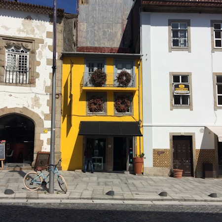 A small, vibrant yellow building with two upper balconies adorned with plants is positioned between two larger older buildings. The adjacent structures have decorative stone details and one has a rustic look, while the other is painted white. A bicycle is parked near a light pole on the pedestrian walkway in front of the yellow building. A sign in one window indicates that the property is for rent.