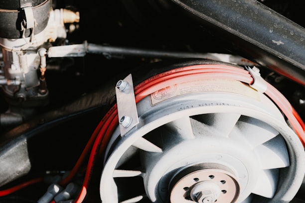 A detailed view of a mechanical component featuring a fan-like structure with visible metallic bolts and nuts. It is surrounded by a loop of red electrical wiring labeled as spark plug wire, set against a background of additional mechanical parts.