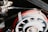 A detailed view of a mechanical component featuring a fan-like structure with visible metallic bolts and nuts. It is surrounded by a loop of red electrical wiring labeled as spark plug wire, set against a background of additional mechanical parts.