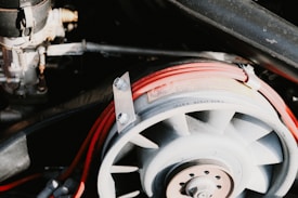 A detailed view of a mechanical component featuring a fan-like structure with visible metallic bolts and nuts. It is surrounded by a loop of red electrical wiring labeled as spark plug wire, set against a background of additional mechanical parts.