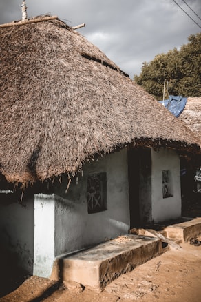 A traditional house with a thatched roof made from natural materials. The walls appear to be made of mud or clay, painted in a light color. The structure includes simple architectural details such as small windows with decorative bars. Surrounding the house, there are indications of a rural setting with trees and utility wires visible in the background.