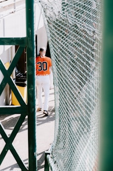 A person wearing a sports uniform with the number 30 on the back is walking away through a narrow space between a fence and a building. The sports uniform consists of a bright orange top and white pants. There is a yellow trash can next to the building on the left side of the person.