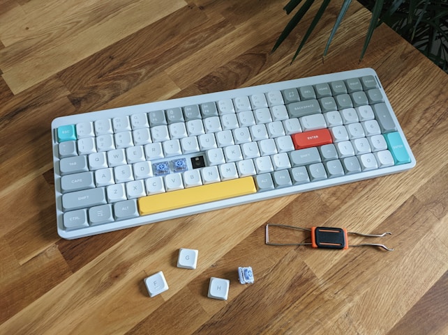 A mechanical keyboard is placed on a wooden surface, featuring a variety of colored keycaps with some keys missing, revealing the switches underneath. Some additional keycaps and a keycap remover tool are positioned nearby.