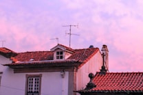 A scenic view of traditional architecture featuring red-tiled rooftops with antennas against a backdrop of a pink and purple sky. The building displays colonial-style design elements, including a prominent chimney and decorative stonework.