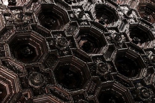 Intricate, geometric woodwork forms a complex pattern of hexagonal shapes, adorned with floral motifs and detailed carvings. The interplay of shadows enhances the depth and richness of the dark wooden material.