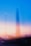 A blurred cityscape features a prominent tall structure in the center against a backdrop of blue and pinkish-orange hues, suggesting a twilight or dawn setting. The lights below form streaks, adding a dynamic and dreamlike quality.