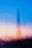 A blurred cityscape features a prominent tall structure in the center against a backdrop of blue and pinkish-orange hues, suggesting a twilight or dawn setting. The lights below form streaks, adding a dynamic and dreamlike quality.