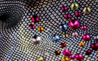 A close-up view of a textured mesh pattern with an array of colorful, scattered beads. The beads vary in size and display a range of colors, including red, pink, blue, silver, and gold. The background features a distorted net-like design, adding depth and movement to the image.