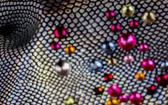 A close-up view of a textured mesh pattern with an array of colorful, scattered beads. The beads vary in size and display a range of colors, including red, pink, blue, silver, and gold. The background features a distorted net-like design, adding depth and movement to the image.