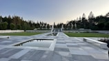 A symmetrical outdoor space featuring a grey tiled pathway leading to a series of steps and a line of vertical pillars surrounded by greenery. The area is bordered by manicured grass and trees, with water features reflecting the sky above.