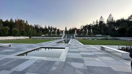 A symmetrical outdoor space featuring a grey tiled pathway leading to a series of steps and a line of vertical pillars surrounded by greenery. The area is bordered by manicured grass and trees, with water features reflecting the sky above.