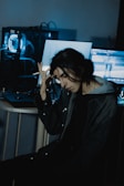 A person with long dark hair sits in a dimly lit room, holding their head with one hand. A computer with blue lighting is visible in the background, along with a couple of screens displaying indistinct content.