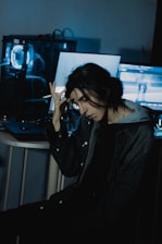 A person with long dark hair sits in a dimly lit room, holding their head with one hand. A computer with blue lighting is visible in the background, along with a couple of screens displaying indistinct content.