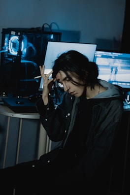 A person with long dark hair sits in a dimly lit room, holding their head with one hand. A computer with blue lighting is visible in the background, along with a couple of screens displaying indistinct content.