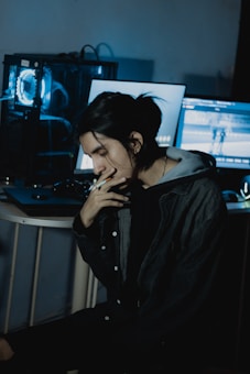 A person with long hair is sitting at a desk, looking thoughtful or contemplative. Two computer monitors are in the background, softly illuminating the scene with blue and cool tones. The setting appears to be a dimly lit room, contributing to a moody atmosphere.