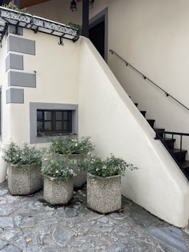 A corner of a building featuring a small outdoor staircase with a black metal railing ascending along a cream-colored wall. Four large, textured concrete planters with green foliage and small flowers are arranged on a stone-paved ground. A window with dark framing is near the bottom of the stairs, framed in gray. Decorative wrought iron elements adorn the roof edge.