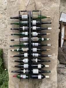 A set of wine bottles is neatly organized in a wall-mounted metal rack against a stone surface. The bottles vary in size and label design, and they are aligned horizontally with the necks pointing outward. The background includes textured stone and patches of greenery, contributing to a rustic and quaint setting.