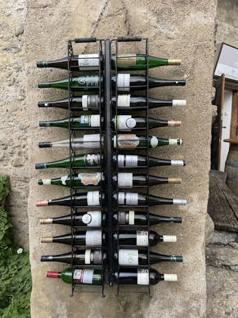 A set of wine bottles is neatly organized in a wall-mounted metal rack against a stone surface. The bottles vary in size and label design, and they are aligned horizontally with the necks pointing outward. The background includes textured stone and patches of greenery, contributing to a rustic and quaint setting.