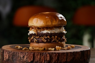 A gourmet burger stacked high with multiple beef patties, crispy fried onions, and a dollop of creamy sauce, all sandwiched between a glossy, toasted bun. The burger is artistically presented on a rustic wooden board with a blurred natural background.