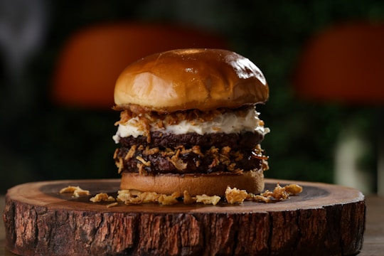 A gourmet burger stacked high with multiple beef patties, crispy fried onions, and a dollop of creamy sauce, all sandwiched between a glossy, toasted bun. The burger is artistically presented on a rustic wooden board with a blurred natural background.