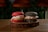 Two gourmet burgers are placed on a round wooden board. One burger bun is red with sesame seeds, filled with a green patty, sliced onions, and other toppings. The other burger bun is black with sesame seeds, containing a brown patty, sliced onions, and similar toppings. The background is blurred, with hints of greenery and warm lighting.