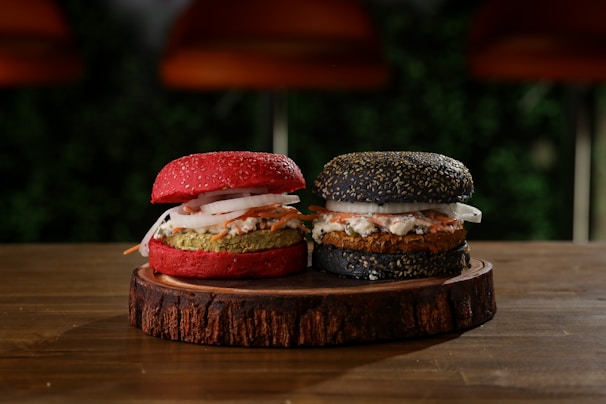 Two gourmet burgers are placed on a round wooden board. One burger bun is red with sesame seeds, filled with a green patty, sliced onions, and other toppings. The other burger bun is black with sesame seeds, containing a brown patty, sliced onions, and similar toppings. The background is blurred, with hints of greenery and warm lighting.