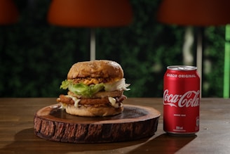 A large, appetizing burger with sesame seed bun, lettuce, fried onion rings, cheese, and a crispy patty, placed on a wooden plate. Next to it is a red can of Coca-Cola, set on a wooden table with a blurred, green leafy background.