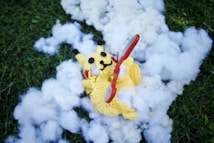 A crocheted yellow character lies on a patch of grass surrounded by fluffy white stuffing. A red and silver pair of scissors is placed across its body.