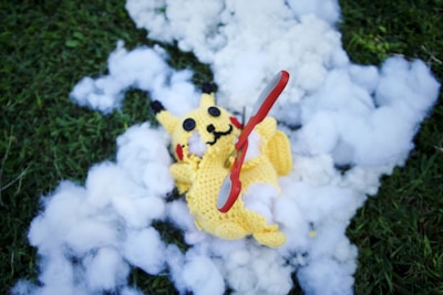 A crocheted yellow character lies on a patch of grass surrounded by fluffy white stuffing. A red and silver pair of scissors is placed across its body.