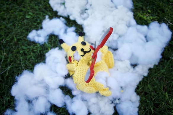 A crocheted yellow character lies on a patch of grass surrounded by fluffy white stuffing. A red and silver pair of scissors is placed across its body.