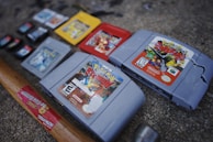 Several video game cartridges, including Nintendo 64 and Game Boy games, are arranged on a rough surface. A hammer with a wooden handle is positioned near the cartridges, and it appears some of the cartridges are cracked or damaged.