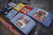 Several video game cartridges, including Nintendo 64 and Game Boy games, are arranged on a rough surface. A hammer with a wooden handle is positioned near the cartridges, and it appears some of the cartridges are cracked or damaged.