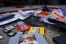 A collection of various Nintendo game cartridges and boxes scattered on the ground. Visible items include cartridges for Game Boy, Game Boy Color, and Nintendo DS, featuring Pokémon titles prominently. There are also some game manuals and packaging materials mixed among the cartridges.