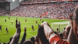 A football match in a large stadium is underway, with players in action on the field. The stands are full of spectators who are cheering and raising their arms, creating a lively and energetic atmosphere. The field is green and well-maintained, and the seating areas are filled with people wearing team colors.