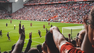 A football match in a large stadium is underway, with players in action on the field. The stands are full of spectators who are cheering and raising their arms, creating a lively and energetic atmosphere. The field is green and well-maintained, and the seating areas are filled with people wearing team colors.