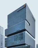 A modern office building with a sleek, glass façade and the AstraZeneca logo prominently displayed near the top. The structure comprises multiple stacked rectangular sections, giving it a distinctive architectural design. Adjacent buildings are also visible in the background, reflecting a cityscape environment.