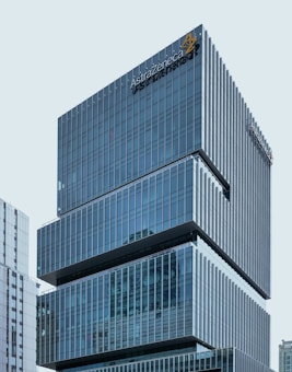 A modern office building with a sleek, glass façade and the AstraZeneca logo prominently displayed near the top. The structure comprises multiple stacked rectangular sections, giving it a distinctive architectural design. Adjacent buildings are also visible in the background, reflecting a cityscape environment.