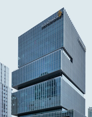 A modern office building with a sleek, glass façade and the AstraZeneca logo prominently displayed near the top. The structure comprises multiple stacked rectangular sections, giving it a distinctive architectural design. Adjacent buildings are also visible in the background, reflecting a cityscape environment.