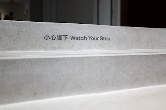 Concrete stairs with a bilingual 'Watch Your Step' warning sign in both Chinese and English. The steps appear clean and slightly worn, situated in an indoor environment with soft lighting.
