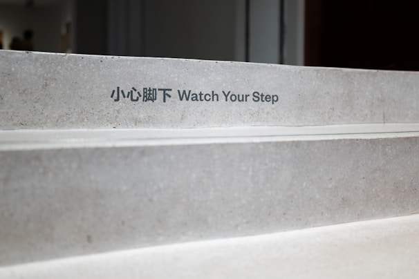 Concrete stairs with a bilingual 'Watch Your Step' warning sign in both Chinese and English. The steps appear clean and slightly worn, situated in an indoor environment with soft lighting.