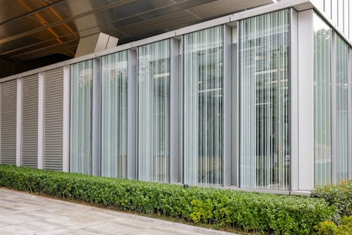 A modern building facade with large vertical glass windows, featuring translucent blinds. A manicured hedge runs along the base of the building, contrasting with the sleek silver and grey exterior.