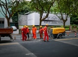 A group of six construction workers in orange uniforms and yellow helmets are working on a road. They are gathered around a hole in the ground, using tools for road maintenance or construction. To the side, a yellow piece of machinery is parked. In the background, there is a large billboard with a smartphone advertisement partially visible. Tall trees line the street and some pedestrians can be seen walking by.