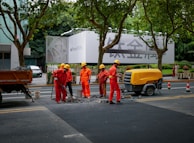 A group of six construction workers in orange uniforms and yellow helmets are working on a road. They are gathered around a hole in the ground, using tools for road maintenance or construction. To the side, a yellow piece of machinery is parked. In the background, there is a large billboard with a smartphone advertisement partially visible. Tall trees line the street and some pedestrians can be seen walking by.