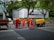 A group of six construction workers in orange uniforms and yellow helmets are working on a road. They are gathered around a hole in the ground, using tools for road maintenance or construction. To the side, a yellow piece of machinery is parked. In the background, there is a large billboard with a smartphone advertisement partially visible. Tall trees line the street and some pedestrians can be seen walking by.