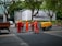 A group of six construction workers in orange uniforms and yellow helmets are working on a road. They are gathered around a hole in the ground, using tools for road maintenance or construction. To the side, a yellow piece of machinery is parked. In the background, there is a large billboard with a smartphone advertisement partially visible. Tall trees line the street and some pedestrians can be seen walking by.