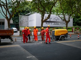 A group of six construction workers in orange uniforms and yellow helmets are working on a road. They are gathered around a hole in the ground, using tools for road maintenance or construction. To the side, a yellow piece of machinery is parked. In the background, there is a large billboard with a smartphone advertisement partially visible. Tall trees line the street and some pedestrians can be seen walking by.
