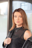 A confident woman wearing a softly draped piece from the Glow From Within collection, standing by a window